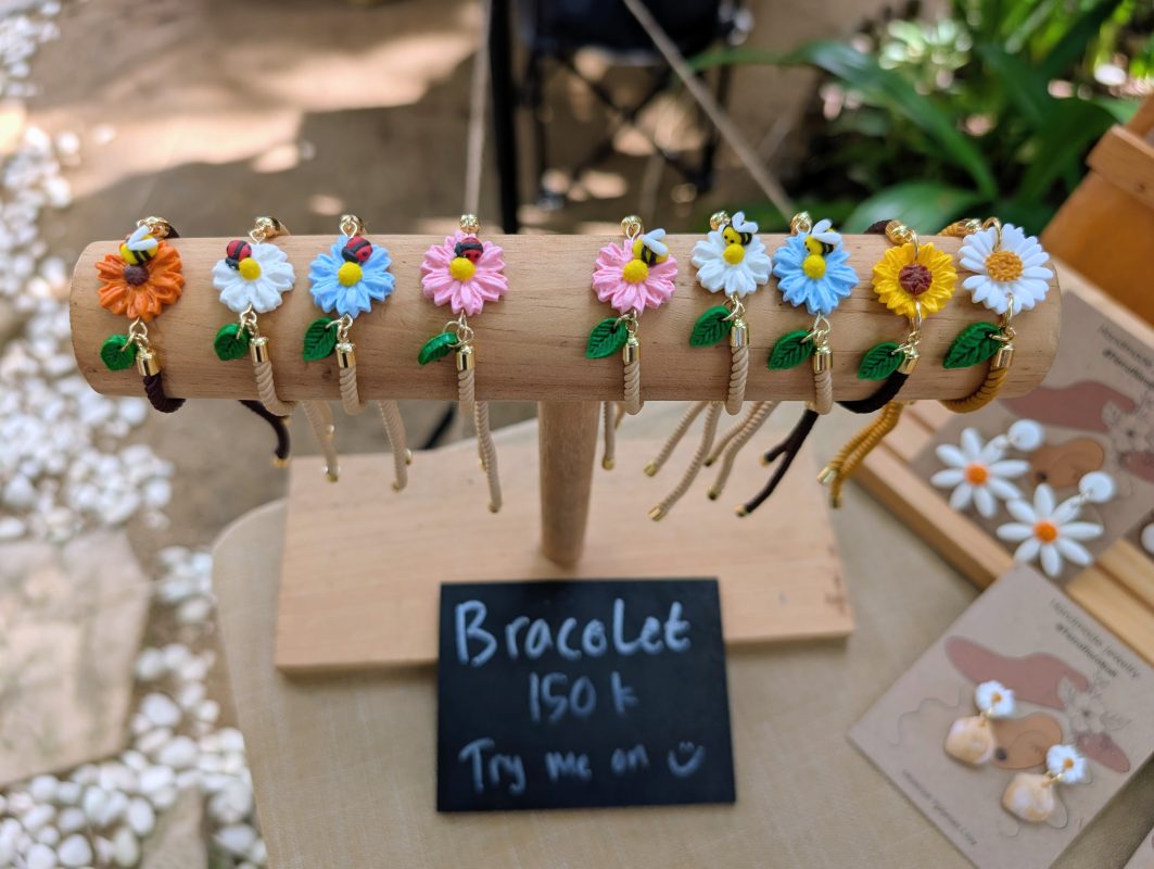 Ten handmade clay flower bracelets with bee and ladybug details on a wooden stand at a Bali market stall