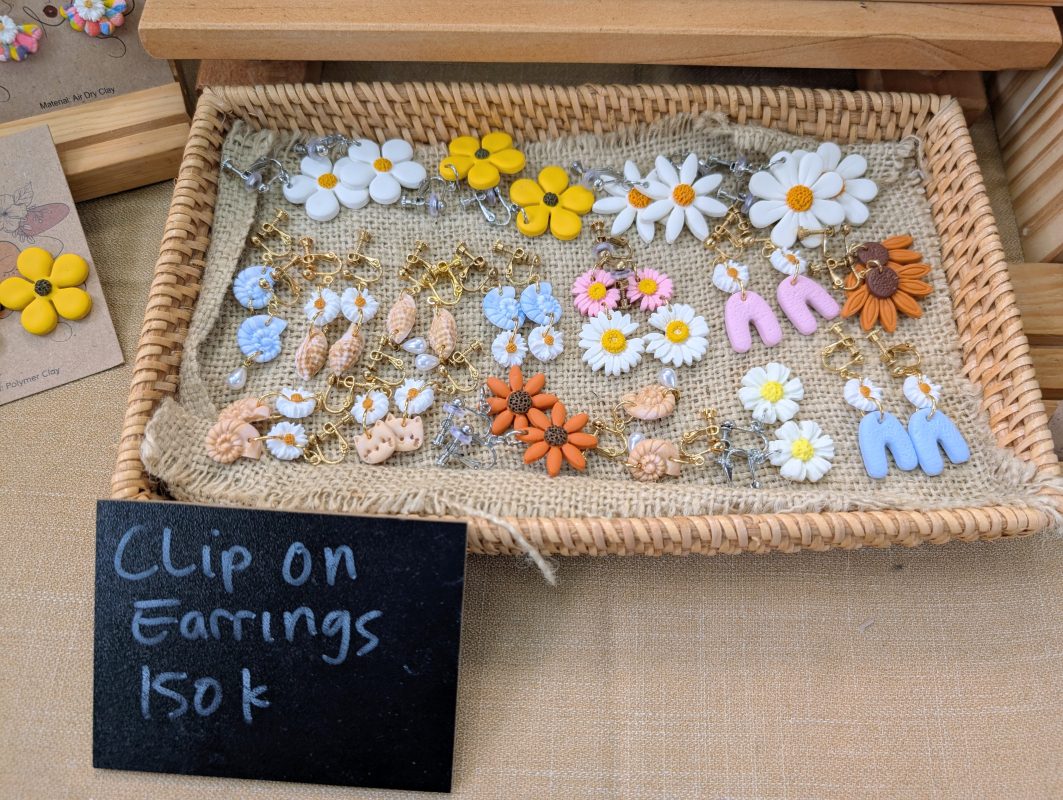 Woven tray of handmade clay clip-on earrings beside a chalkboard sign reading Clip On Earrings 150k
